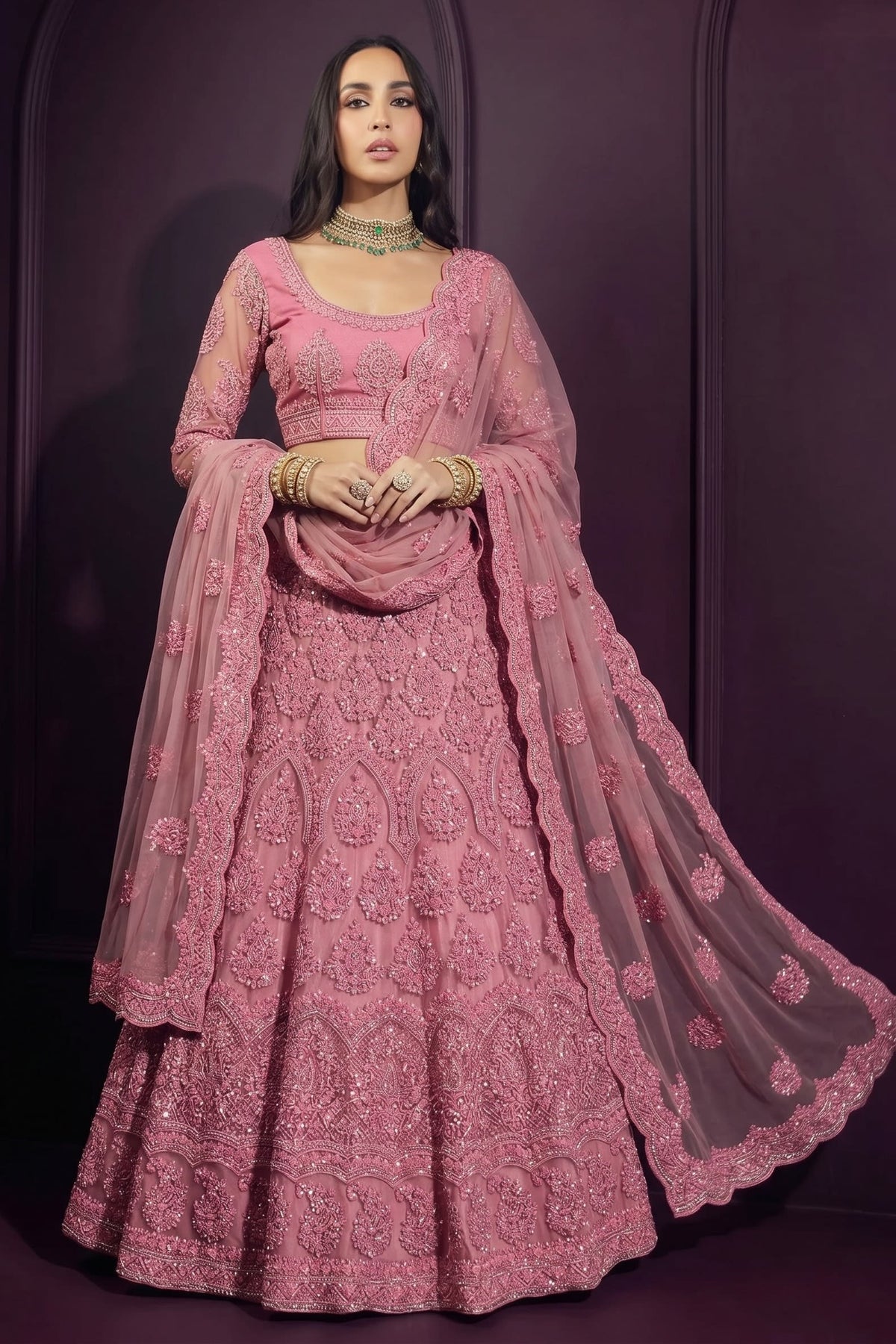 Model in a coral pink wedding guest lehenga with a long-sleeve embroidered choli, flared net skirt and flowing sheer dupatta.