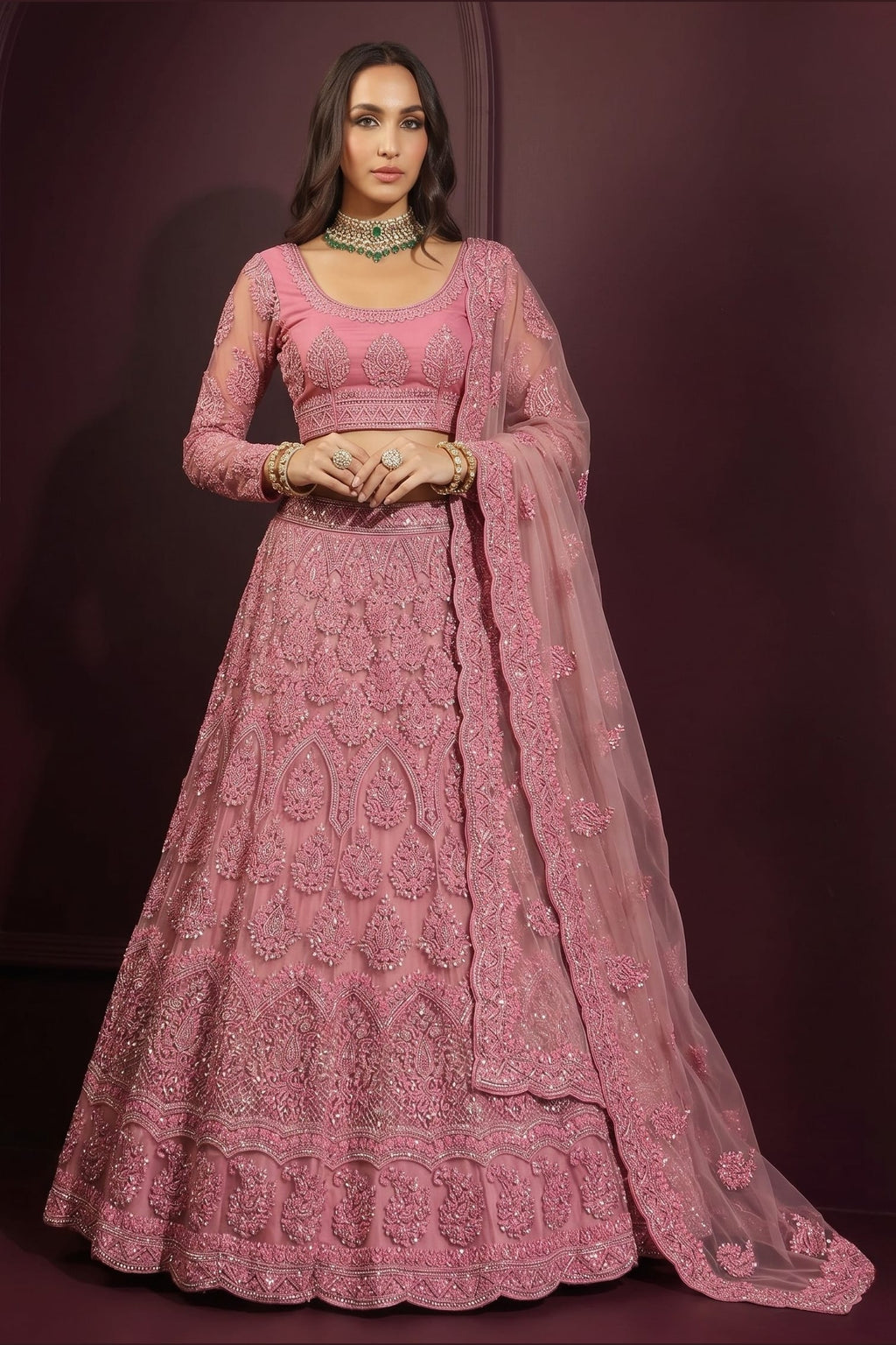 Front view of an indian lehenga choli in coral pink net with dense embroidery, long-sleeve choli and sheer dupatta on one shoulder.