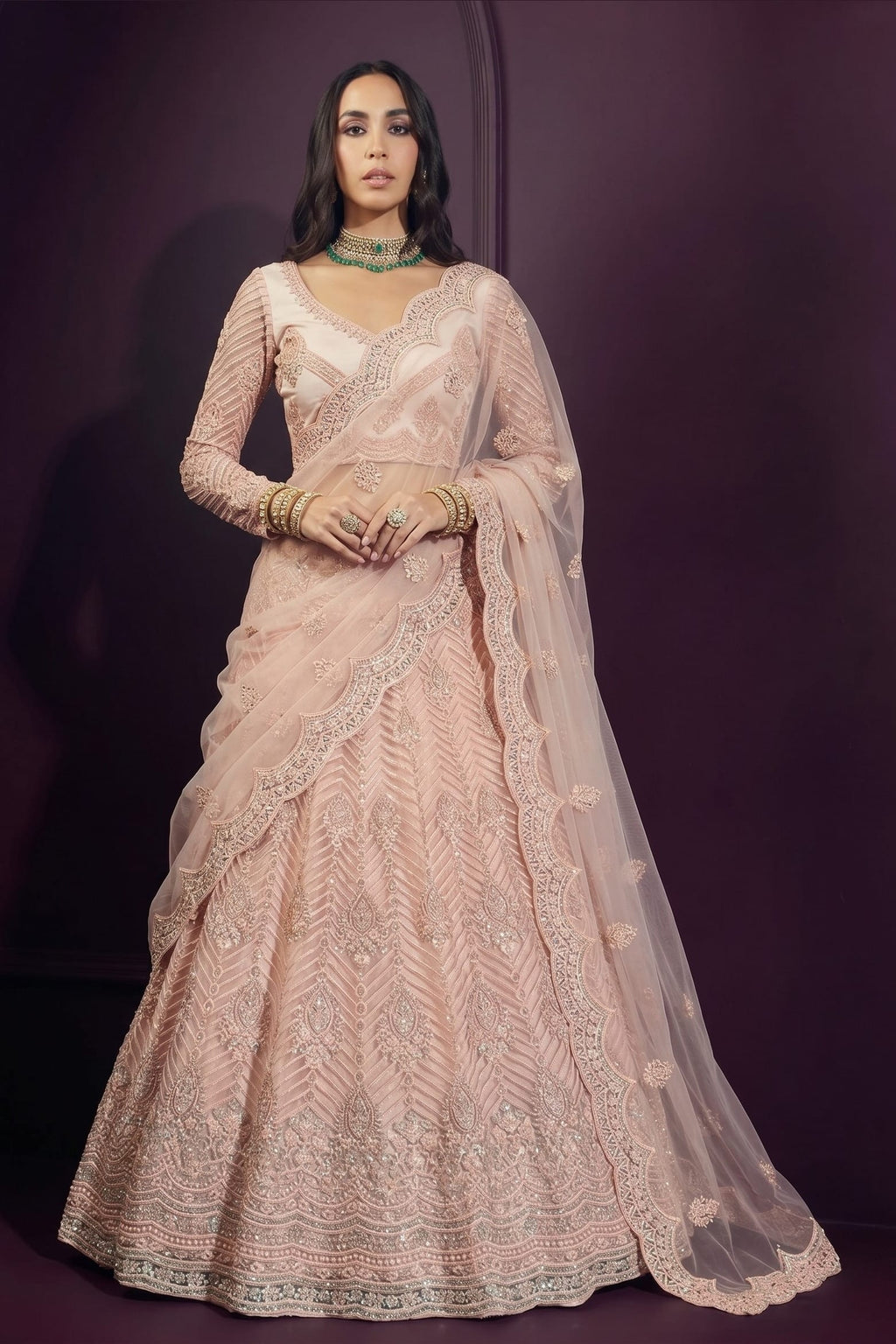Model wearing a pale peach sangeet lehenga choli with a long-sleeve embroidered blouse, flared net skirt and sheer dupatta on one shoulder.