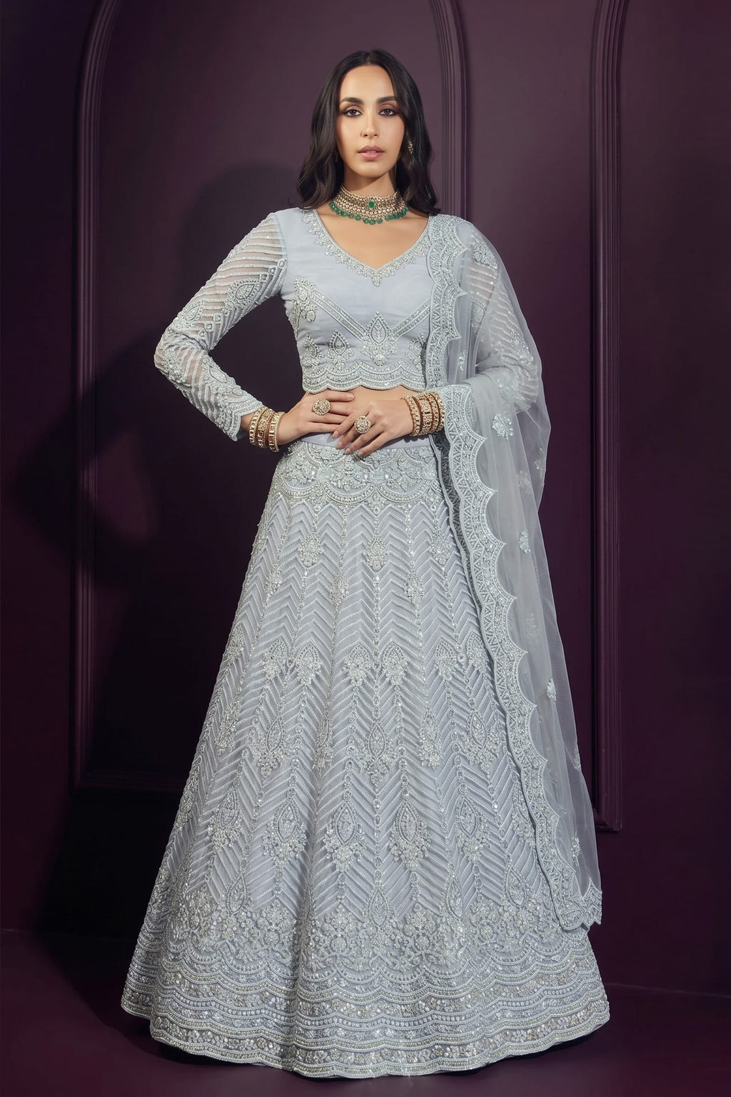 Model in a bluish grey wedding guest lehenga with long-sleeve choli and sheer embroidered dupatta, hands on waist, full front pose.