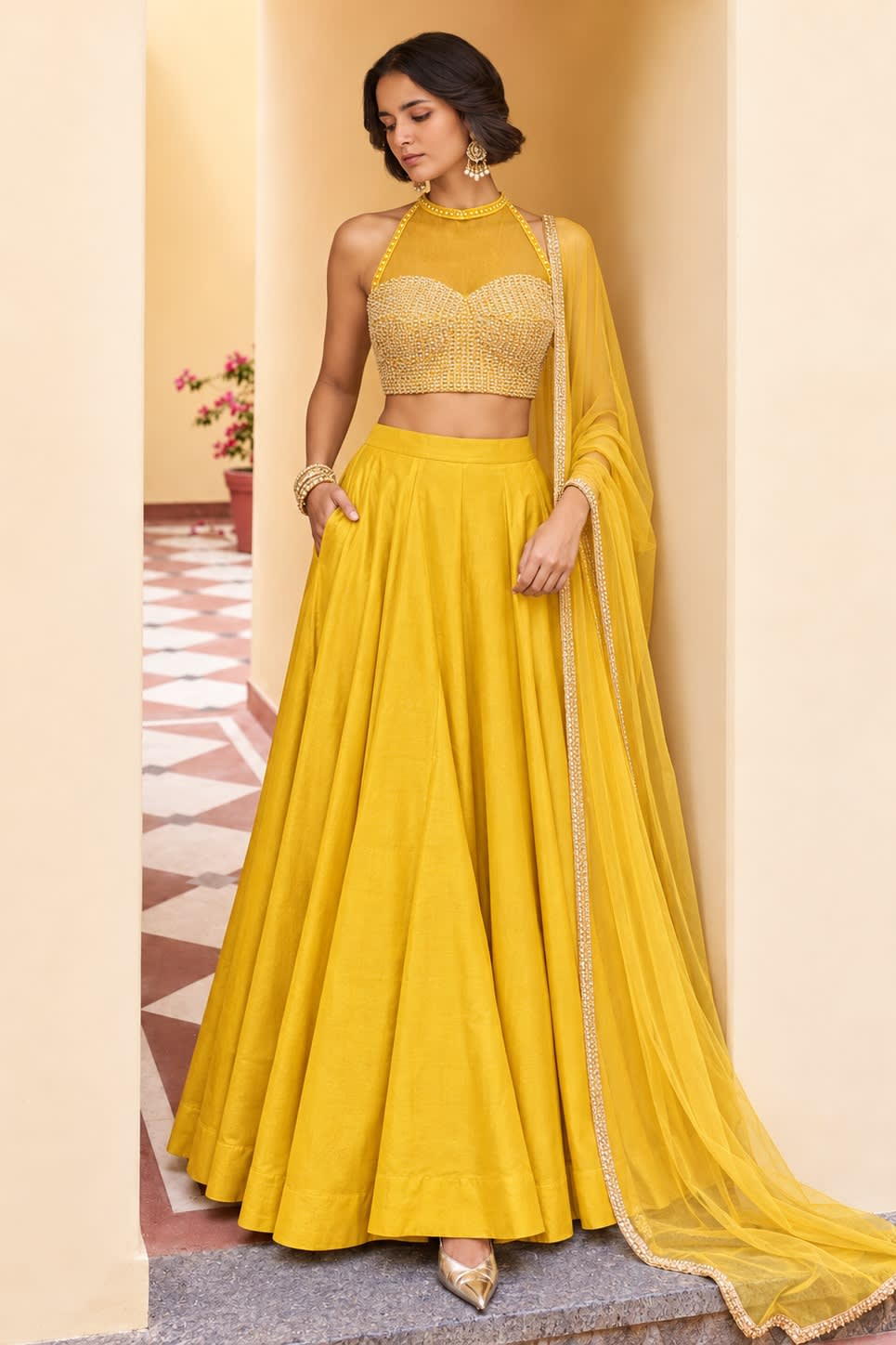 Woman wearing a mustard yellow haldi lehenga in roman silk with embroidered halter net choli and net dupatta, full-length front view.