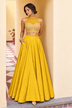 Woman wearing a mustard yellow silk lehenga choli in roman silk with embroidered halter net choli, full-length front view without dupatta.