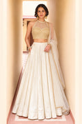 Woman wearing a cream wedding lehenga in roman silk with gold embroidered halter net choli and sheer net dupatta, full-length front view.