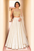 Woman wearing a cream wedding lehenga in roman silk with gold embroidered halter net choli and sheer net dupatta, full-length front view.