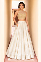Woman wearing a cream lehenga dress style lehenga choli in roman silk with gold embroidered halter net choli, full-length front view.