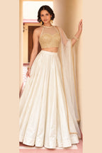 Woman wearing a cream pakistani lehenga in roman silk with embroidered halter net choli and net dupatta with gold border, front view.