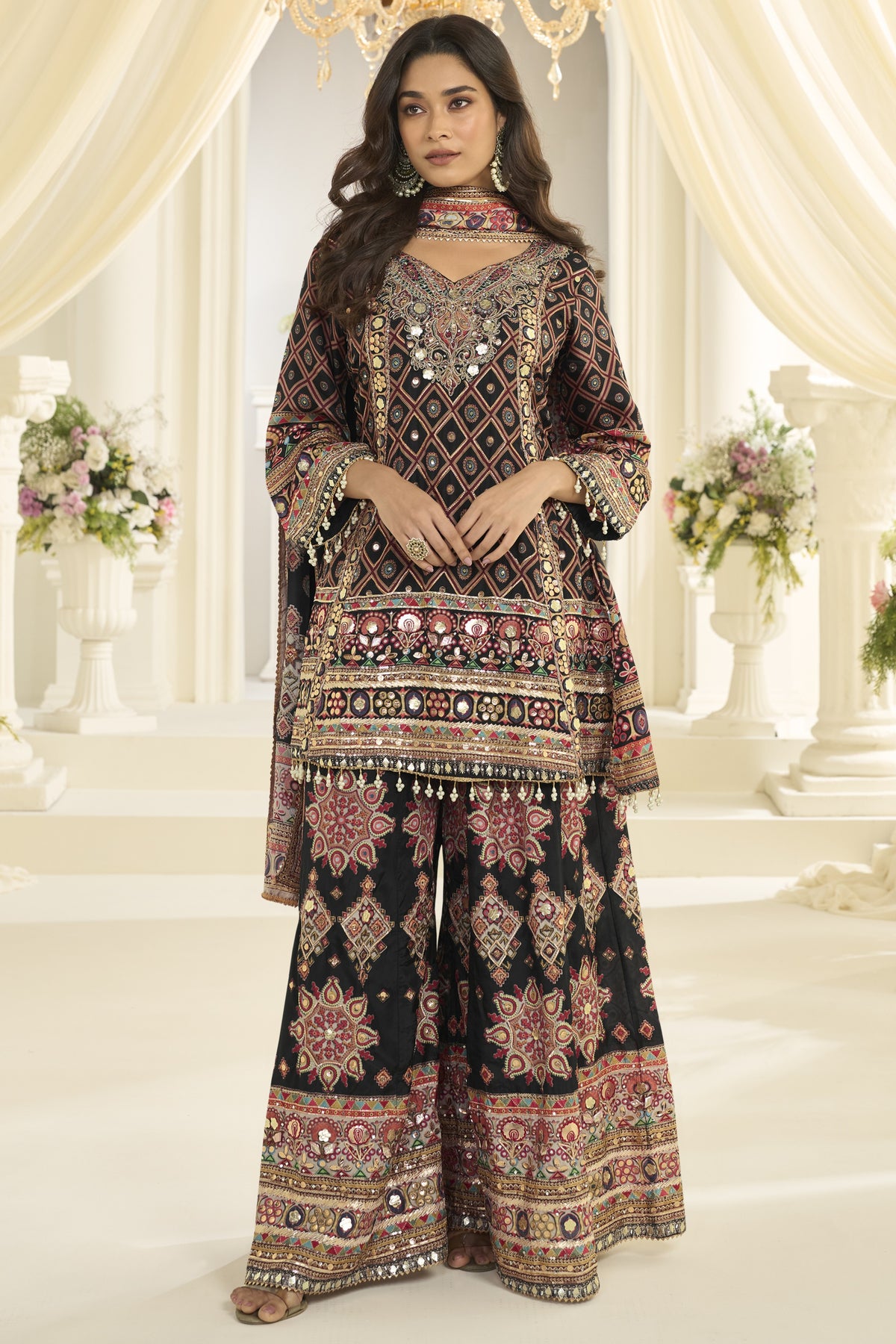 A woman models a traditional South Asian salwar kameez suit set with intricate mirror work and embroidery. The outfit features a black base with detailed multicolor patterns and reflective embellishments, set against a decorated indoor backdrop with draped cream curtains and floral arrangements.