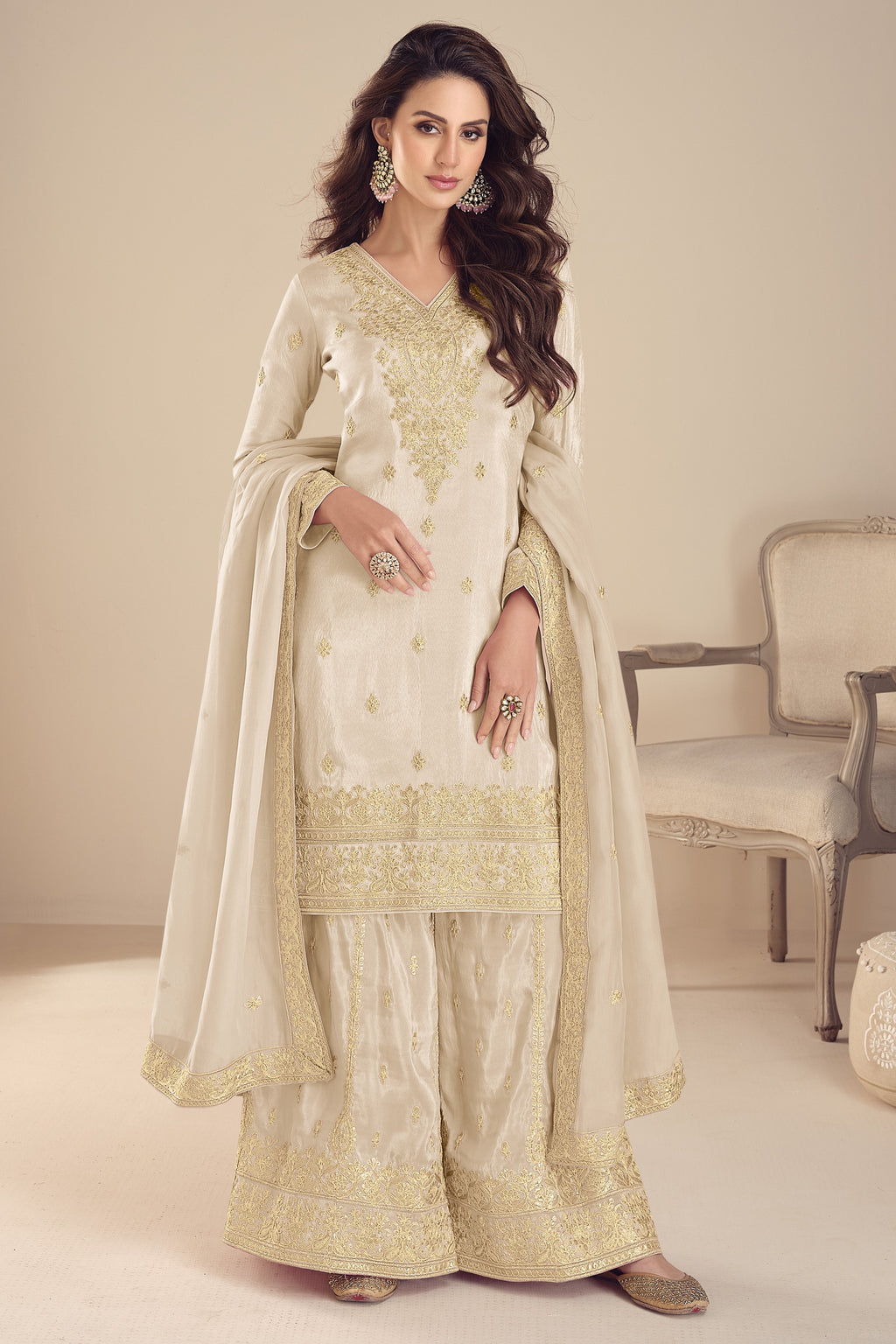 Woman wearing a cream and gold embroidered traditional outfit in a room with a chair.