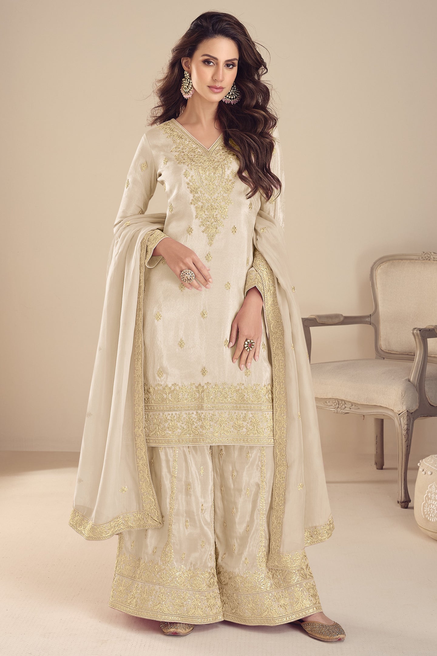 Woman wearing a cream and gold embroidered traditional outfit in a room with a chair.