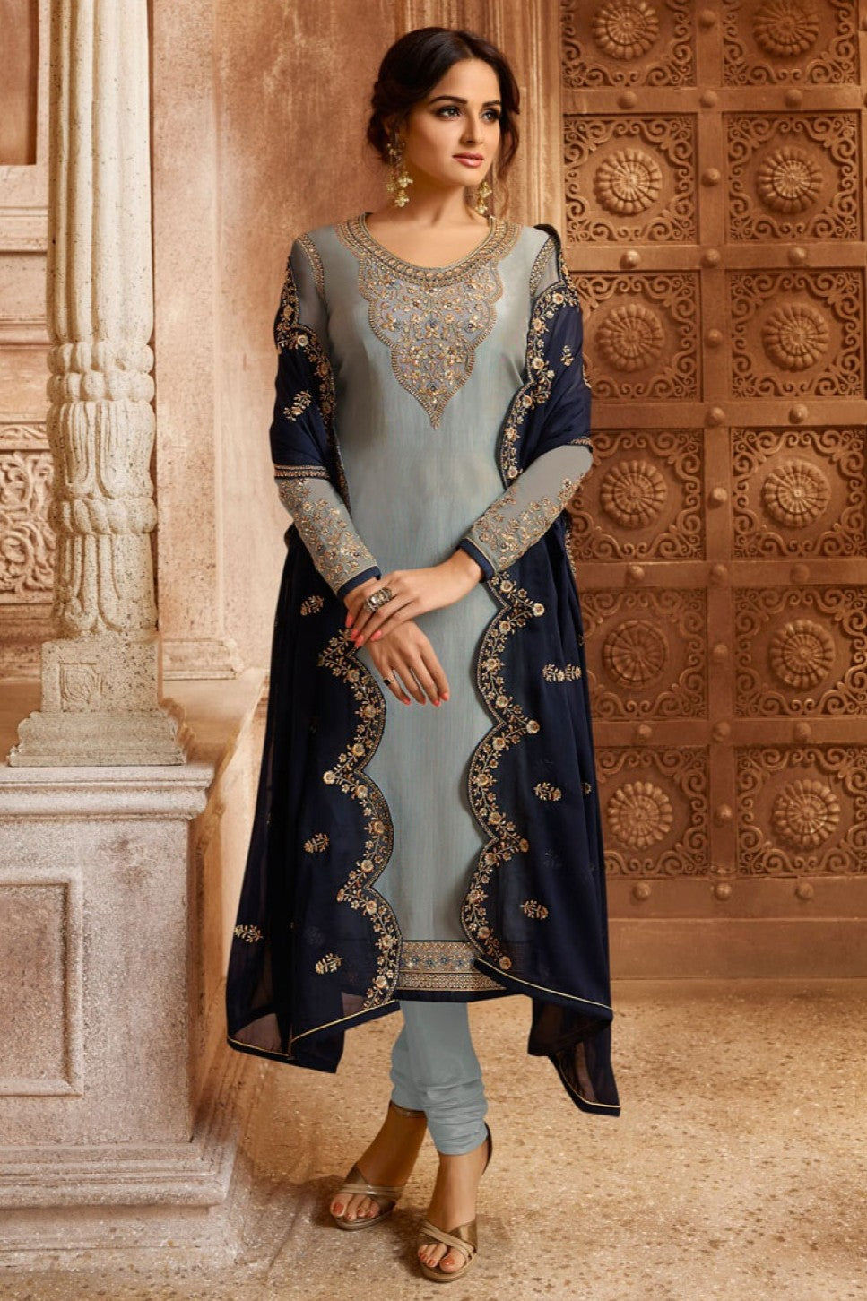 A woman standing in a full front view wearing a light gray kameez for women with a gold embroidered neckline, santoon churidar and a navy dupatta with a scalloped border.