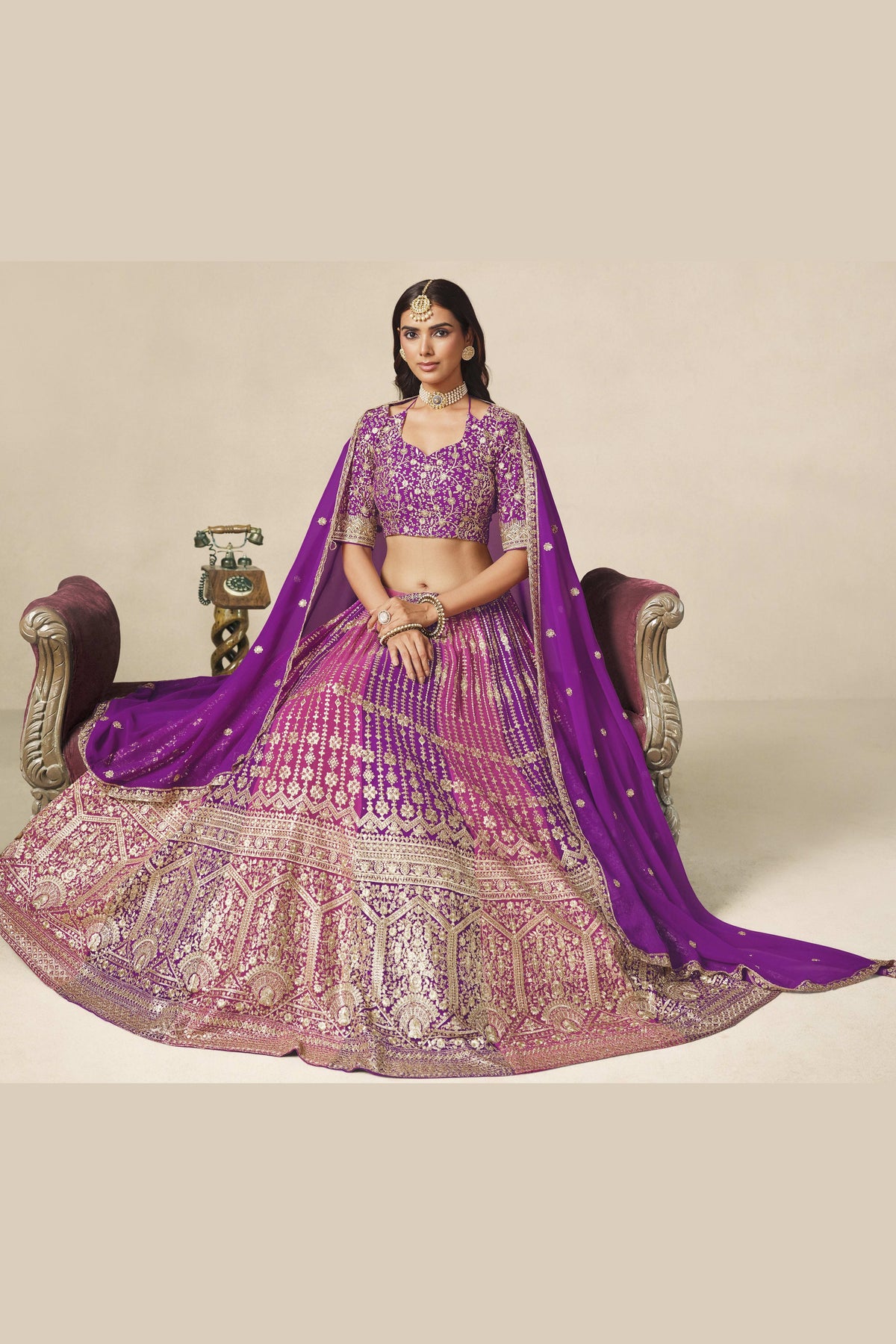 Full-length seated view of a purple indian wedding guest dresses style lehenga in georgette with zari and sequin work on the flared skirt.