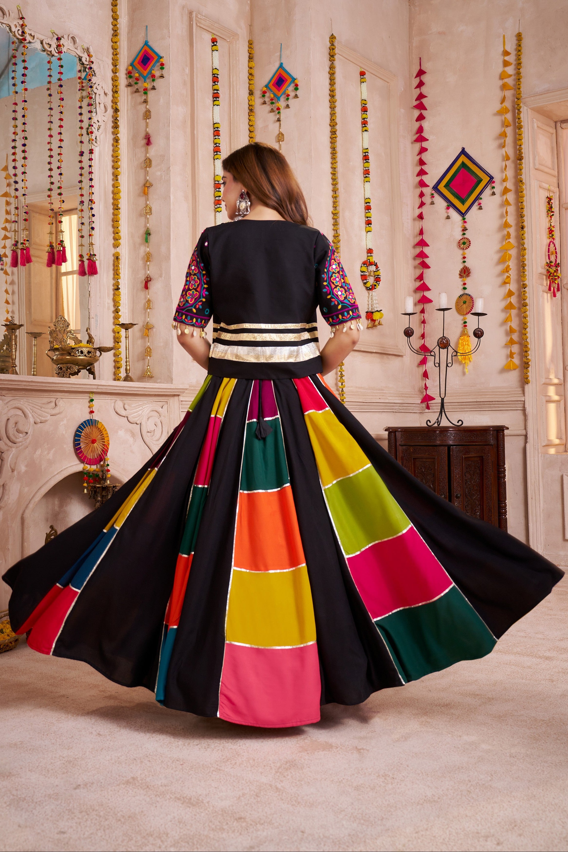 Woman in a colorful traditional outfit with a black base, standing in a decorated room.
