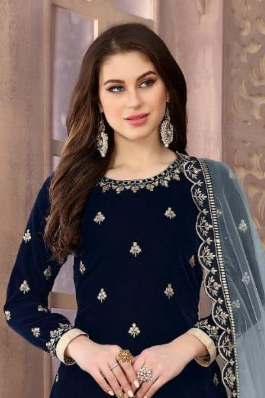 A waist-up view of a woman standing beside a flower urn in a navy blue female punjabi suit with an embroidered velvet kameez and grey net dupatta.