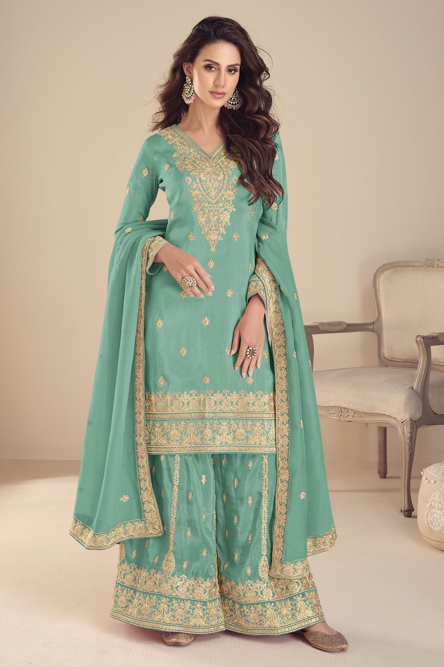 Woman wearing a turquoise traditional outfit with gold embroidery in a neutral setting.