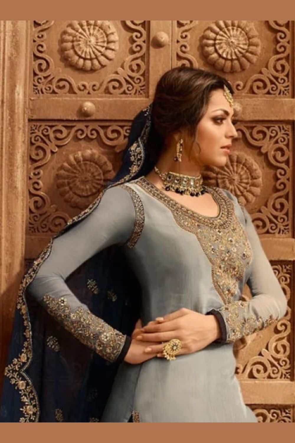 A waist-up side view of a woman with clasped hands wearing light gray pakistani suits with a gold embroidered neckline panel, jhumka earrings and a navy dupatta.