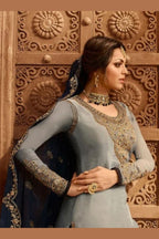 A waist-up side view of a woman with clasped hands wearing light gray pakistani suits with a gold embroidered neckline panel, jhumka earrings and a navy dupatta.