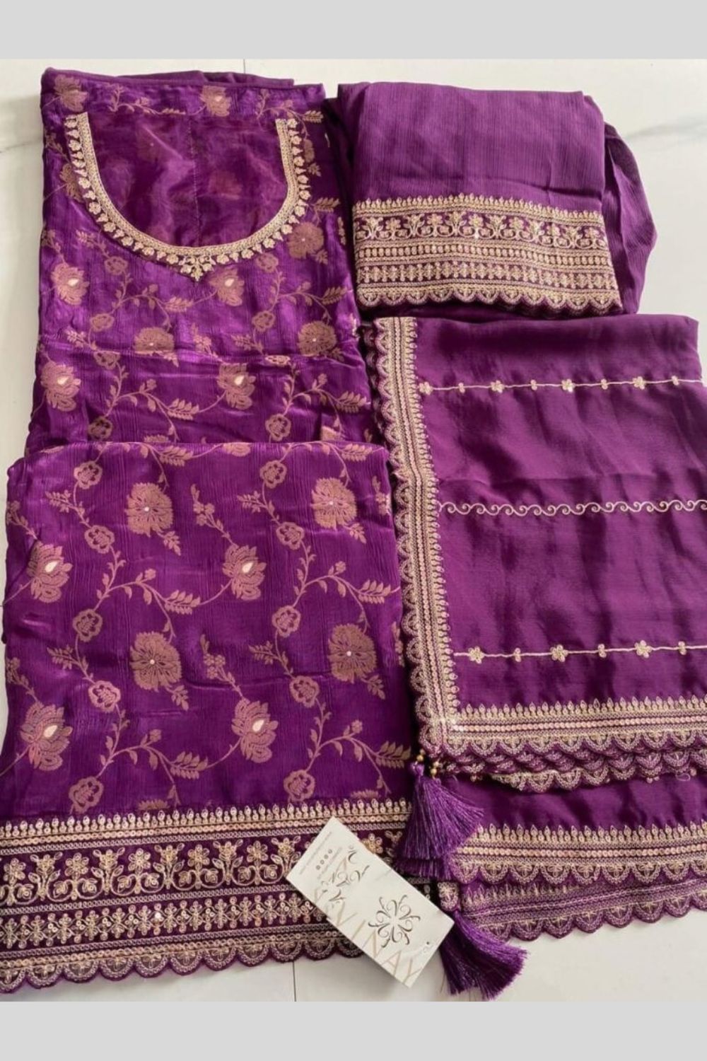 A folded purple ladies plazo suit set with floral jacquard patterns and embroidery, including a kurta, dupatta, and matching fabric pieces, displayed on a white surface.