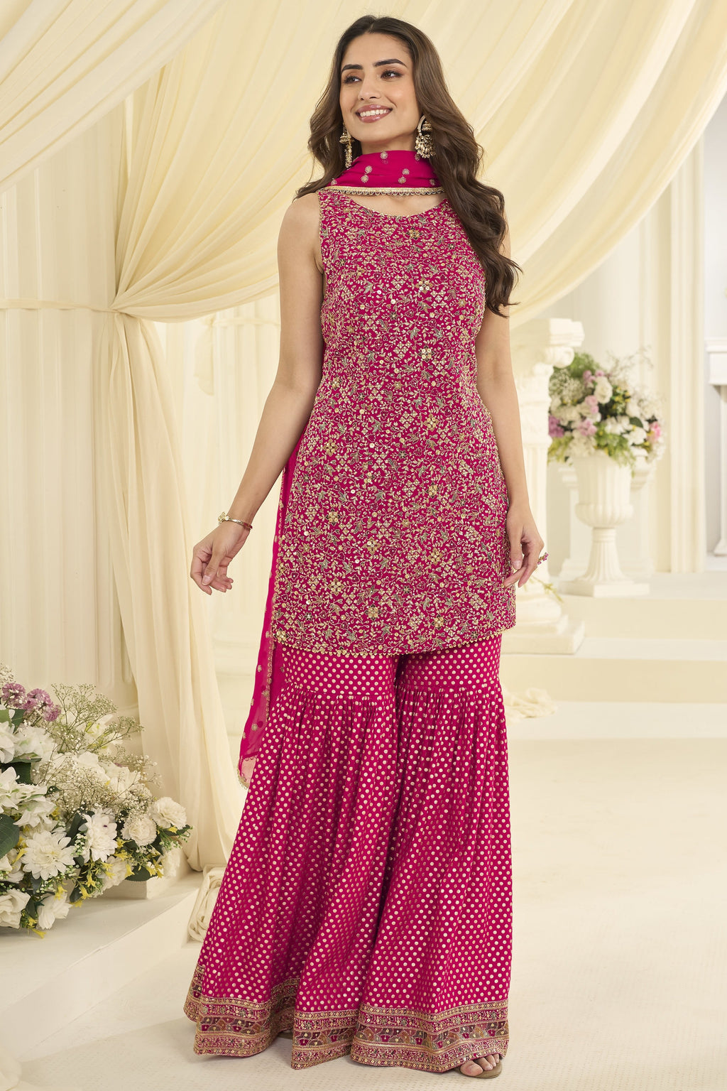 Woman wearing a reddish pink gharara suit in georgette with Swarovski, Zarkan and embroidery work, paired with a woven jacquard gharara, full-length front view.
