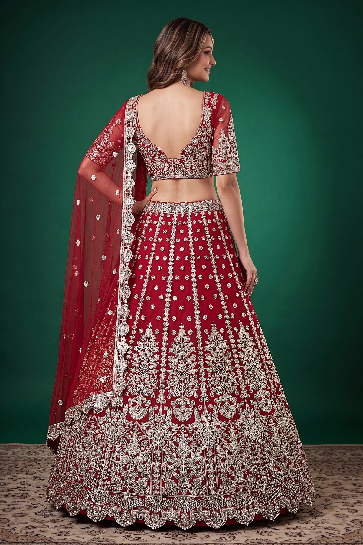Seated view of a red engagement lehenga in net with zari, dori and sequins work throughout on the sleeveless blouse and lehenga.