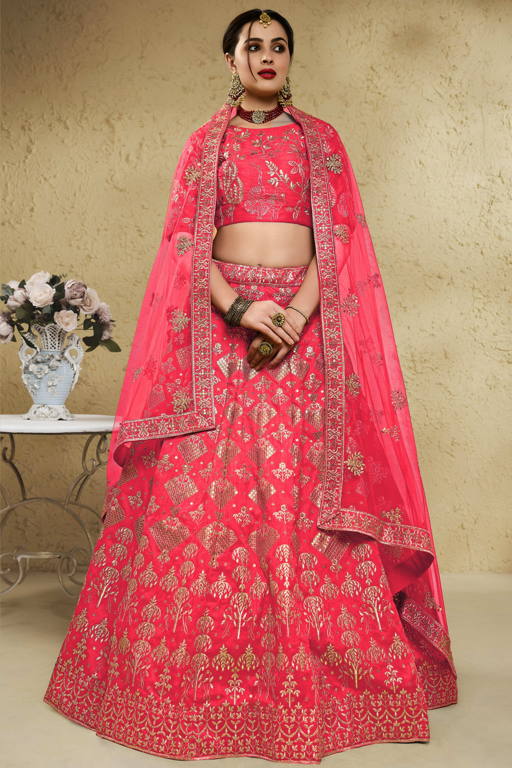 Woman wearing a pinkish red lehenga for wedding in art silk with zari and sequin embroidery, half sleeve boat neck blouse, full-length front view.