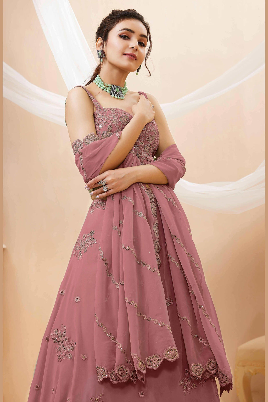 Seated view of a rust pink engagement lehenga in georgette with zari, dori and sequin work on the sleeveless blouse and lehenga.