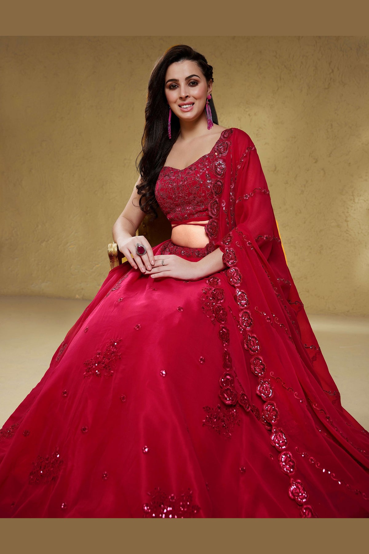 Seated view of a reddish pink party wear lehenga in georgette with zari, dori and sequin work on the sleeveless blouse and lehenga.