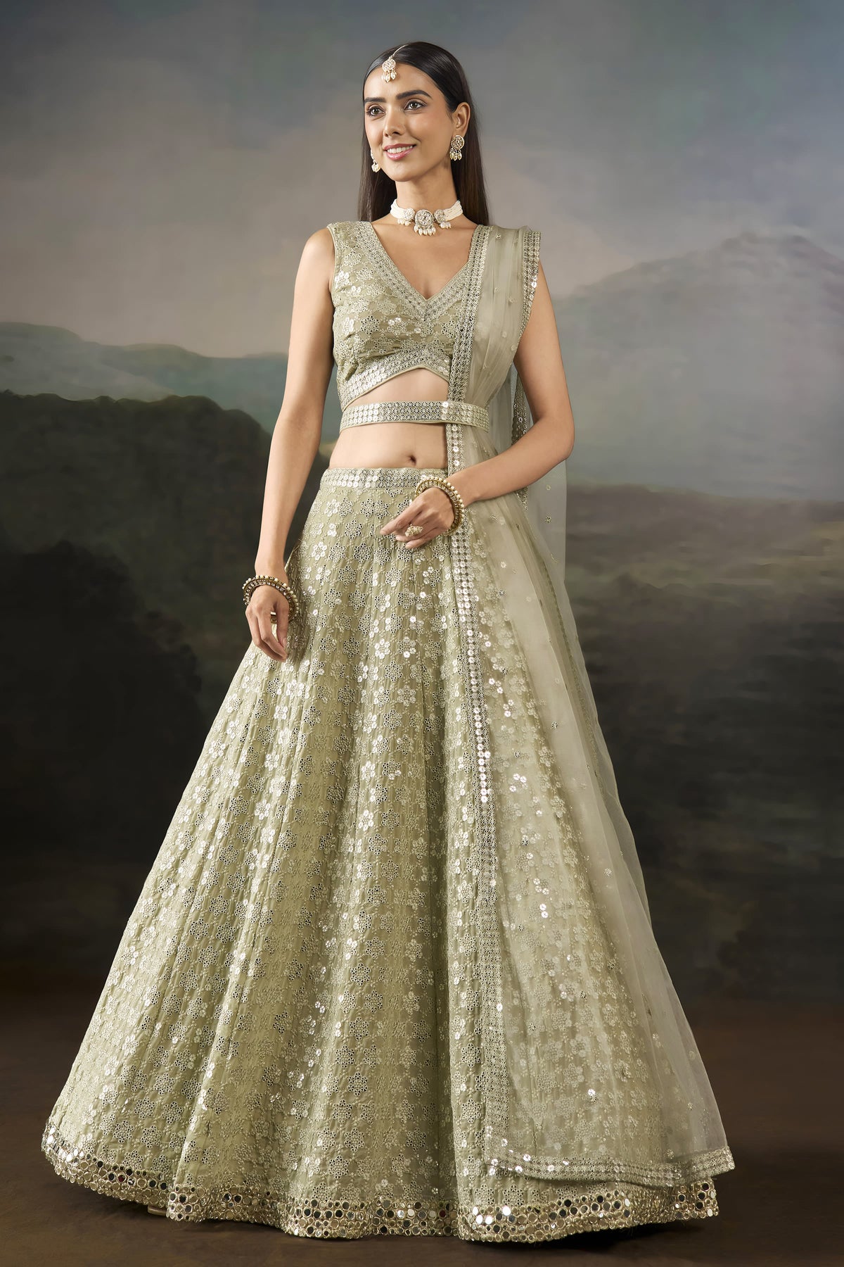 Model wearing a champagne georgette lehenga for mehendi with a sleeveless V neck blouse, worked belt and sheer net dupatta, standing in a full front pose.