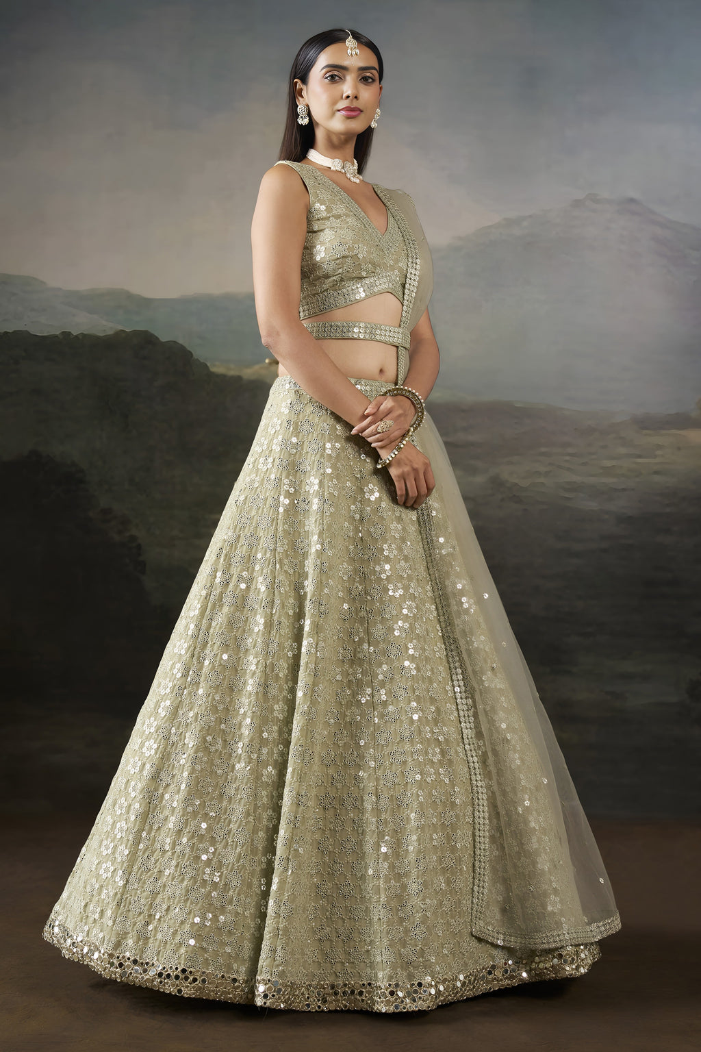 Front pose of the champagne mehndi lehenga with a sleeveless V neck blouse, mirror work belt and long net dupatta over one shoulder.