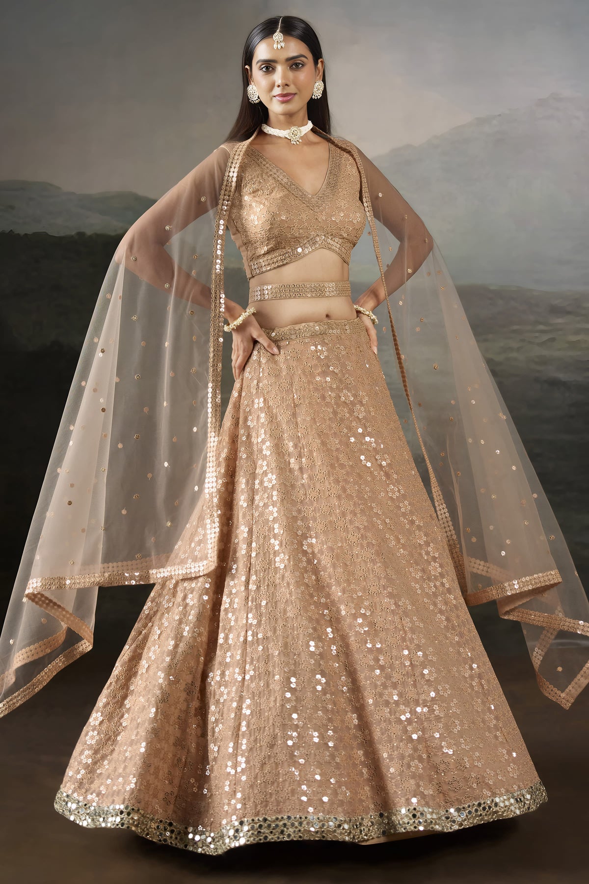 Full front view of the pale peach pastel lehenga with hands on the waist, sleeveless blouse and net dupatta draped over both shoulders.