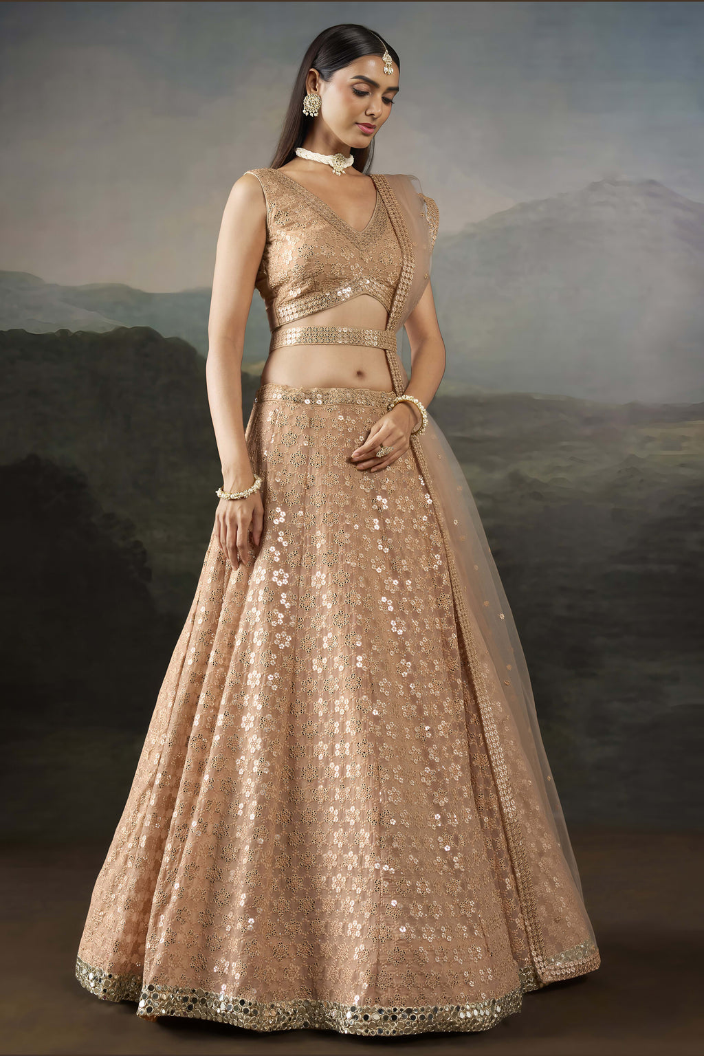 Model in a pale peach pastel lehenga standing front-facing with a sleeveless V neck blouse, worked belt and long net dupatta.