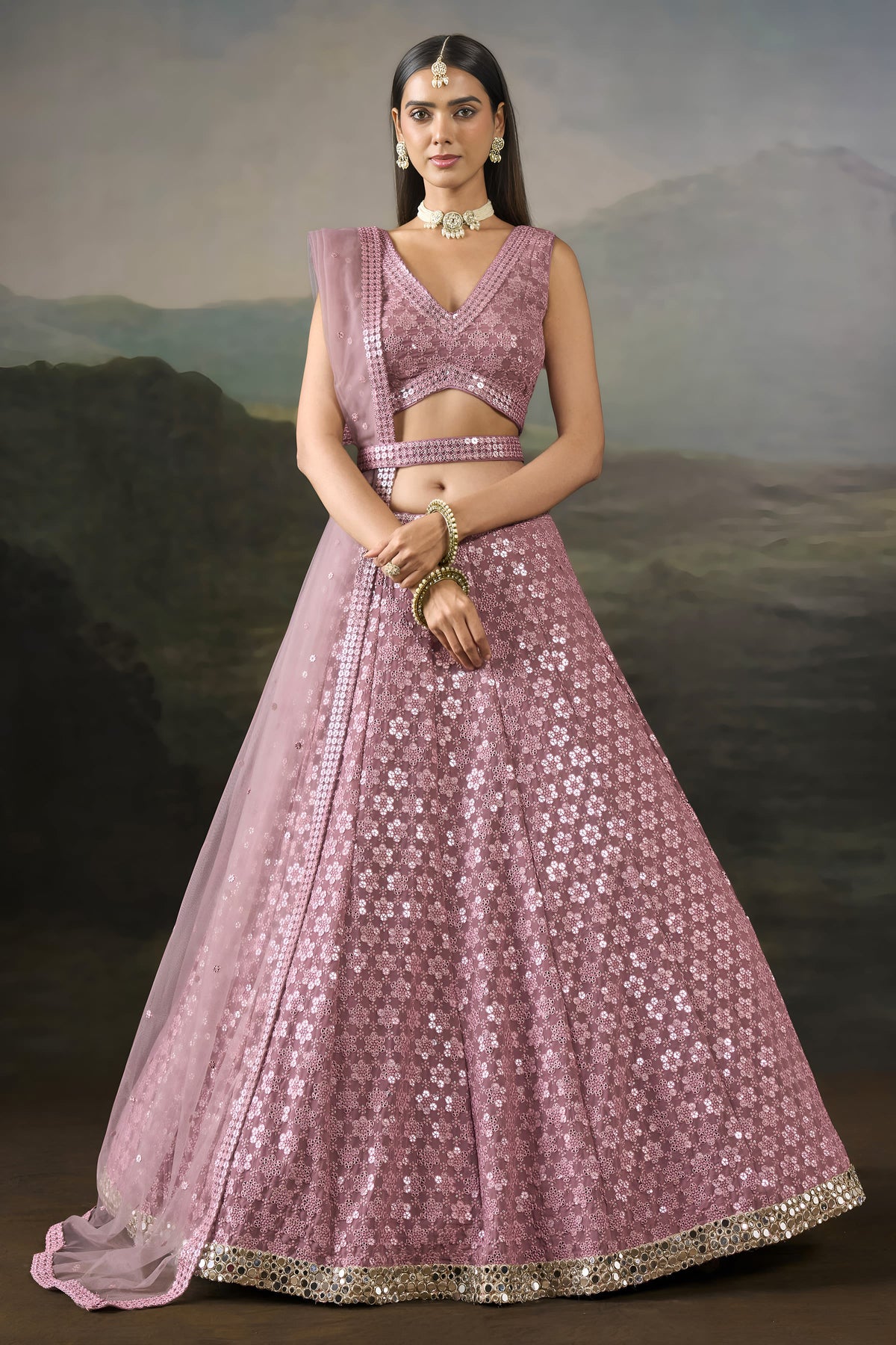 Model wearing a baby pink sangeet lehengas set with a sleeveless V neck blouse, worked belt and sheer net dupatta, standing front-facing.