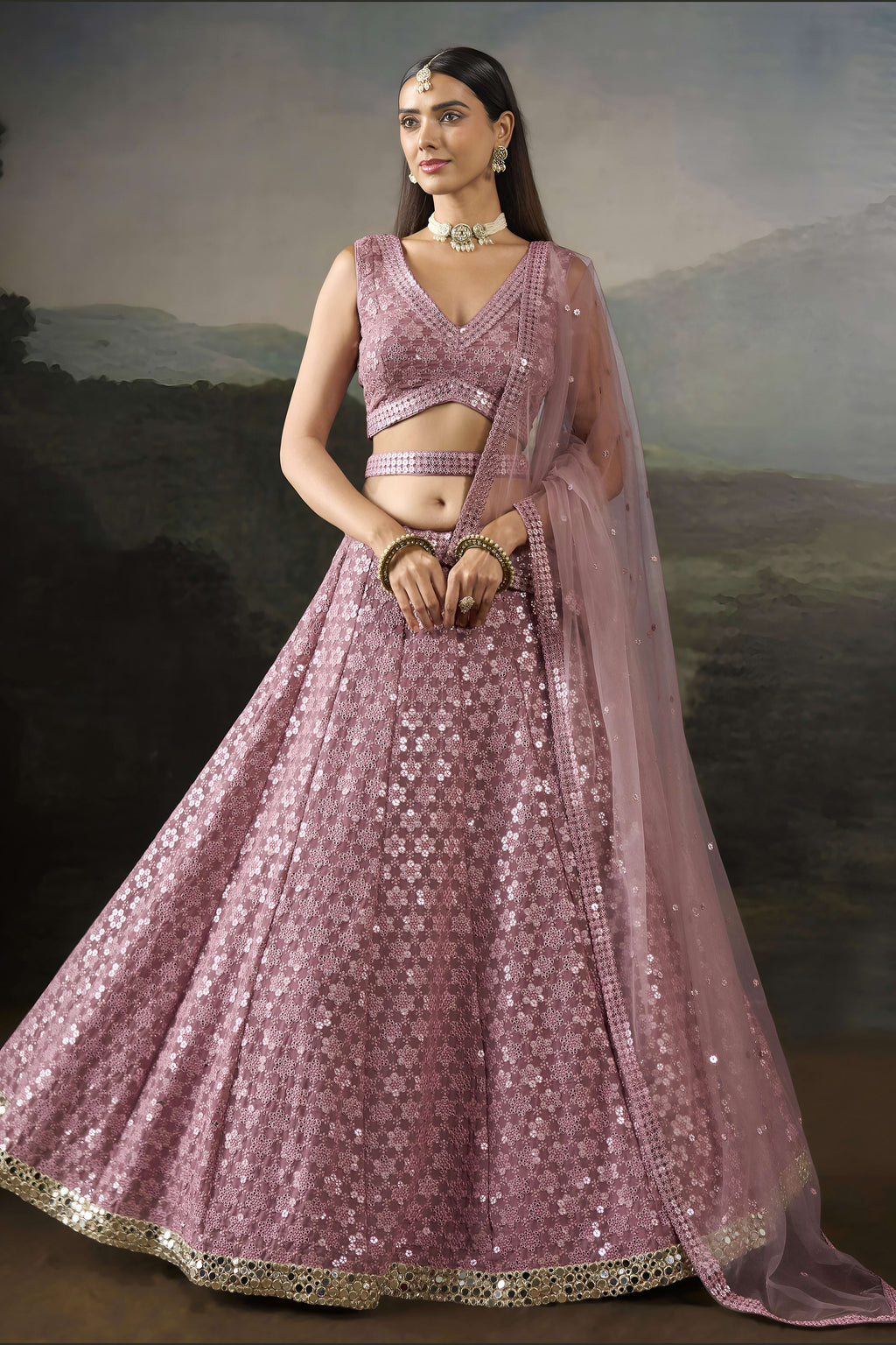 Full front view of the baby pink wedding wear lehenga choli with hands clasped, sleeveless blouse and net dupatta over one shoulder.