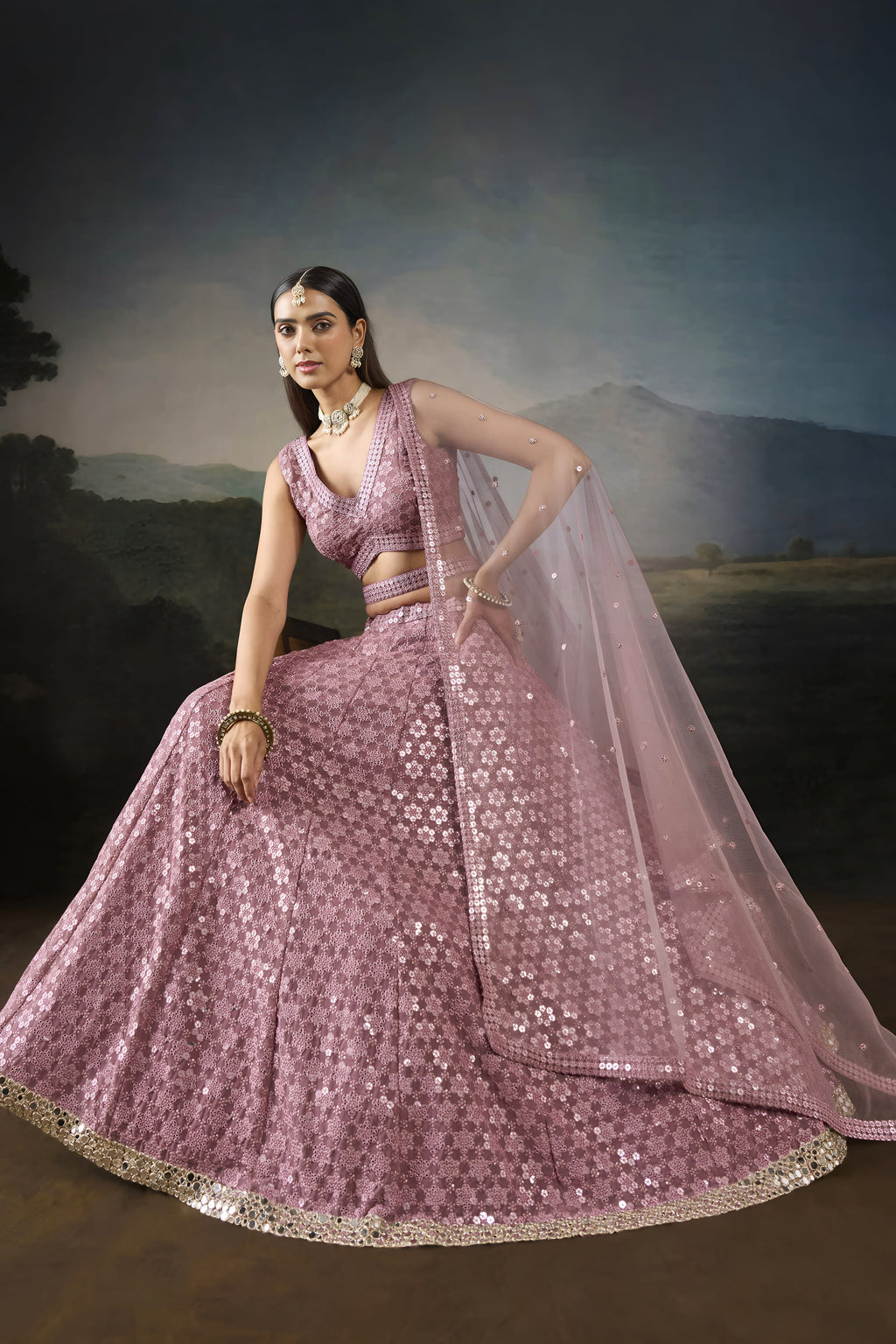 Seated pose of the baby pink sangeet attire with the flared sequinned lehenga spread out and the net dupatta draped to the side.