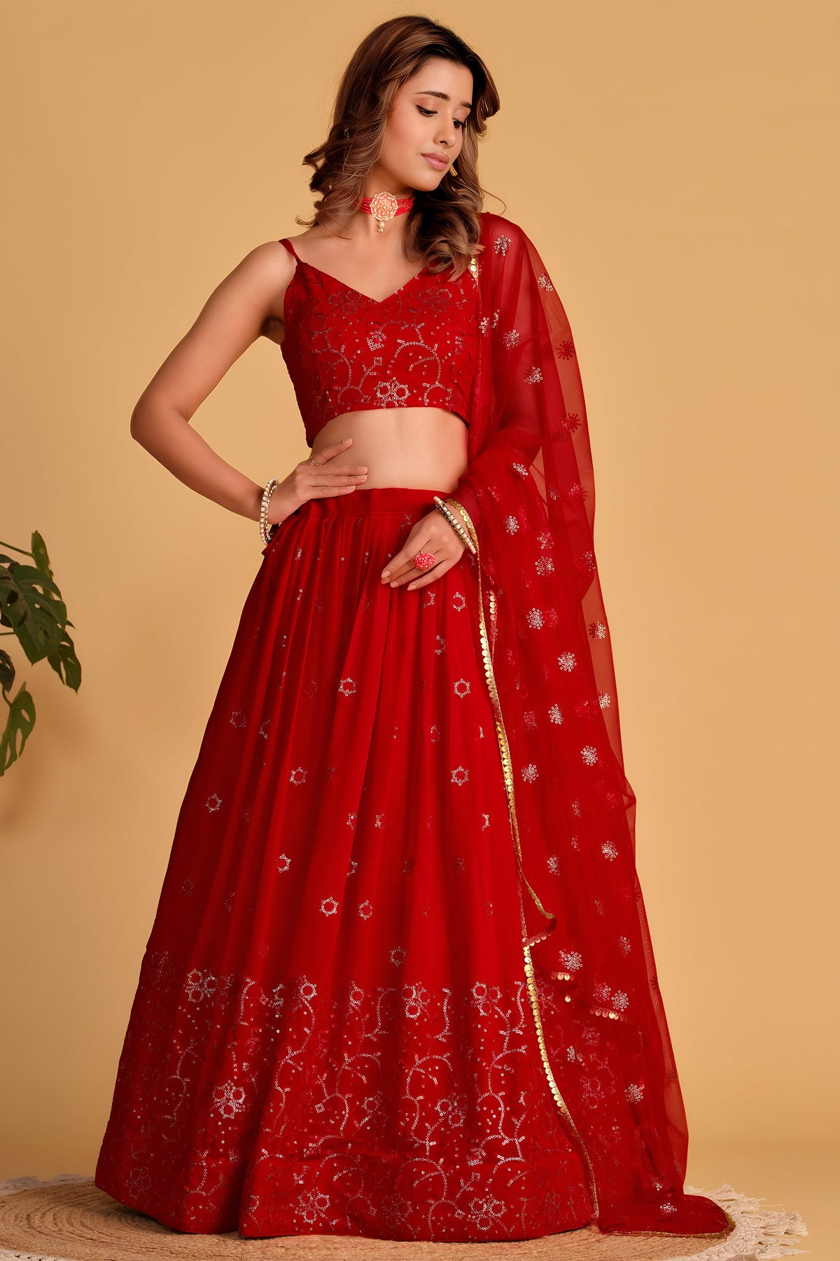 Woman wearing a reddish pink engagement lehenga with zari and sequin embroidery, sleeveless V neck choli, full-length front view.