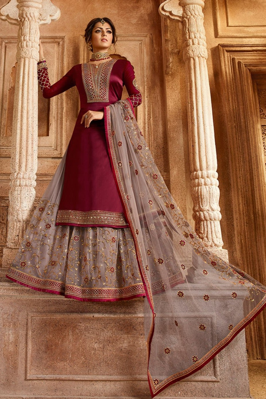 A woman poses in a full-length maroon kameez suit with an embroidered gray dupatta against an ornate architectural backdrop.