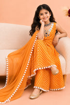 A young girl wearing a orange traditional Indian outfit with white stripes, a matching dupatta with white pom-pom trim, and a decorative mirror work neckline, sitting on a beige sofa against a peach background.