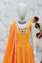 An orange sleeveless dress with white tie-dye stripes and a matching orange dupatta, displayed on a white mannequin. The dress features circular mirror work embellishments on the bodice and a white pom-pom trim along the neckline.